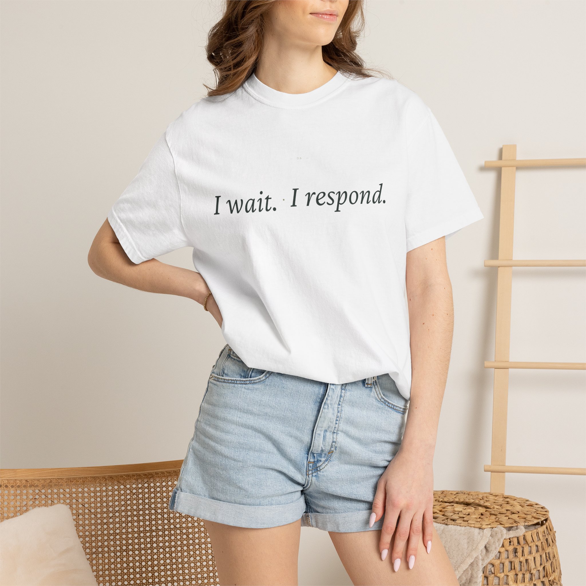 Person wearing a white T-shirt with the text 'I wait. I respond.' and light denim shorts, seated beside wicker furniture in a neutral room.