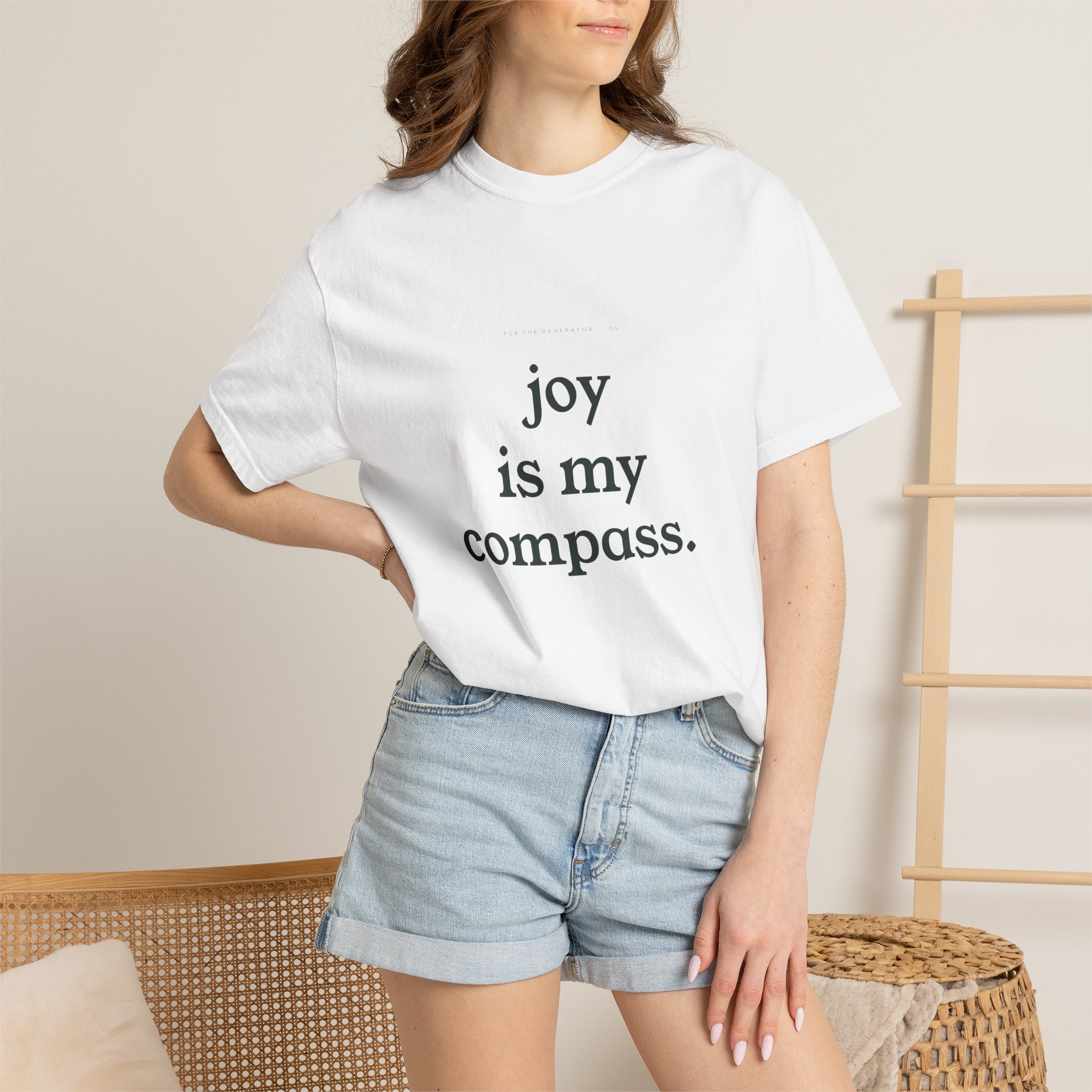 Person wearing a white T-shirt with the text 'joy is my compass' visible, seated beside a woven wicker chair in a neutral room.