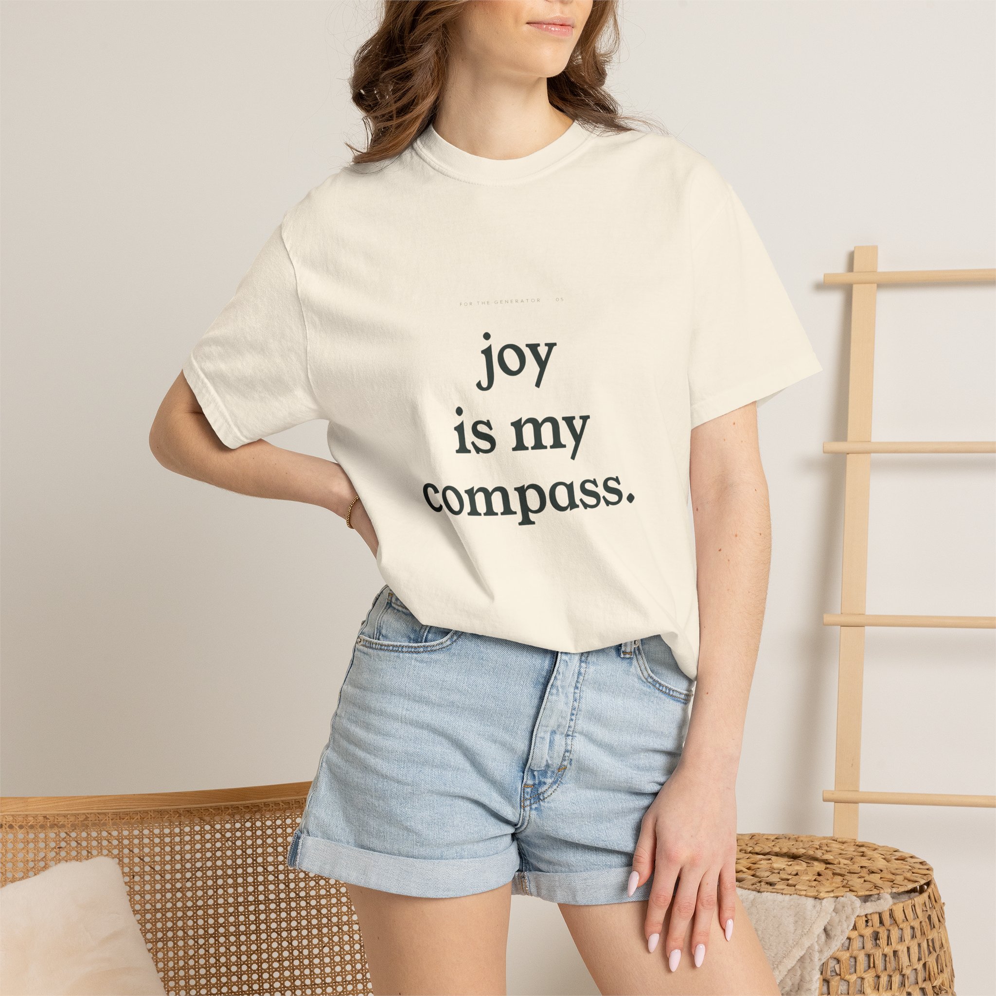 Woman wearing a white T-shirt with the text 'joy is my compass' visible, standing beside a woven chair.